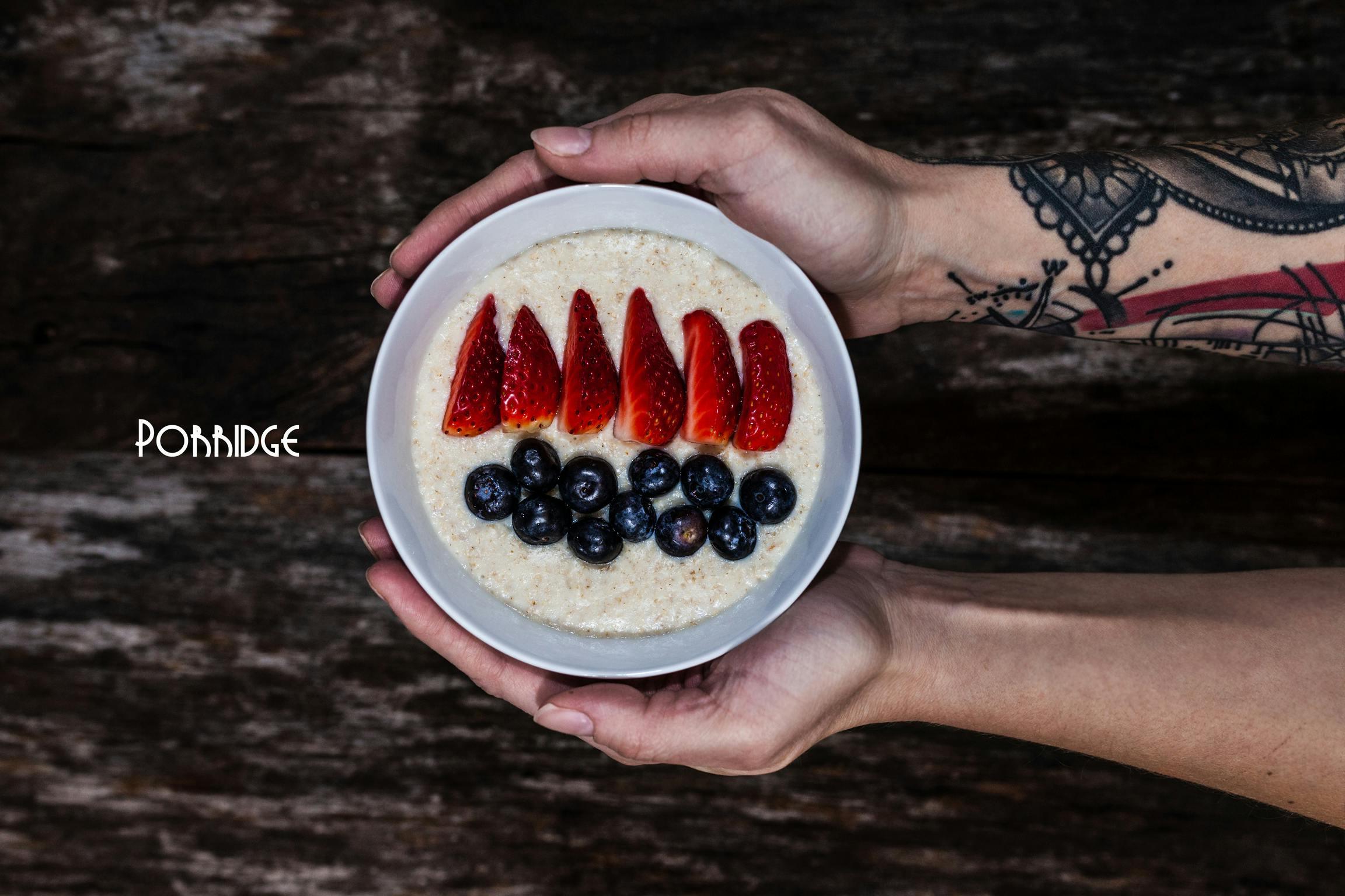 Tattooed hands holding a bowl with porridge and fresh fruits in front of a wooden background