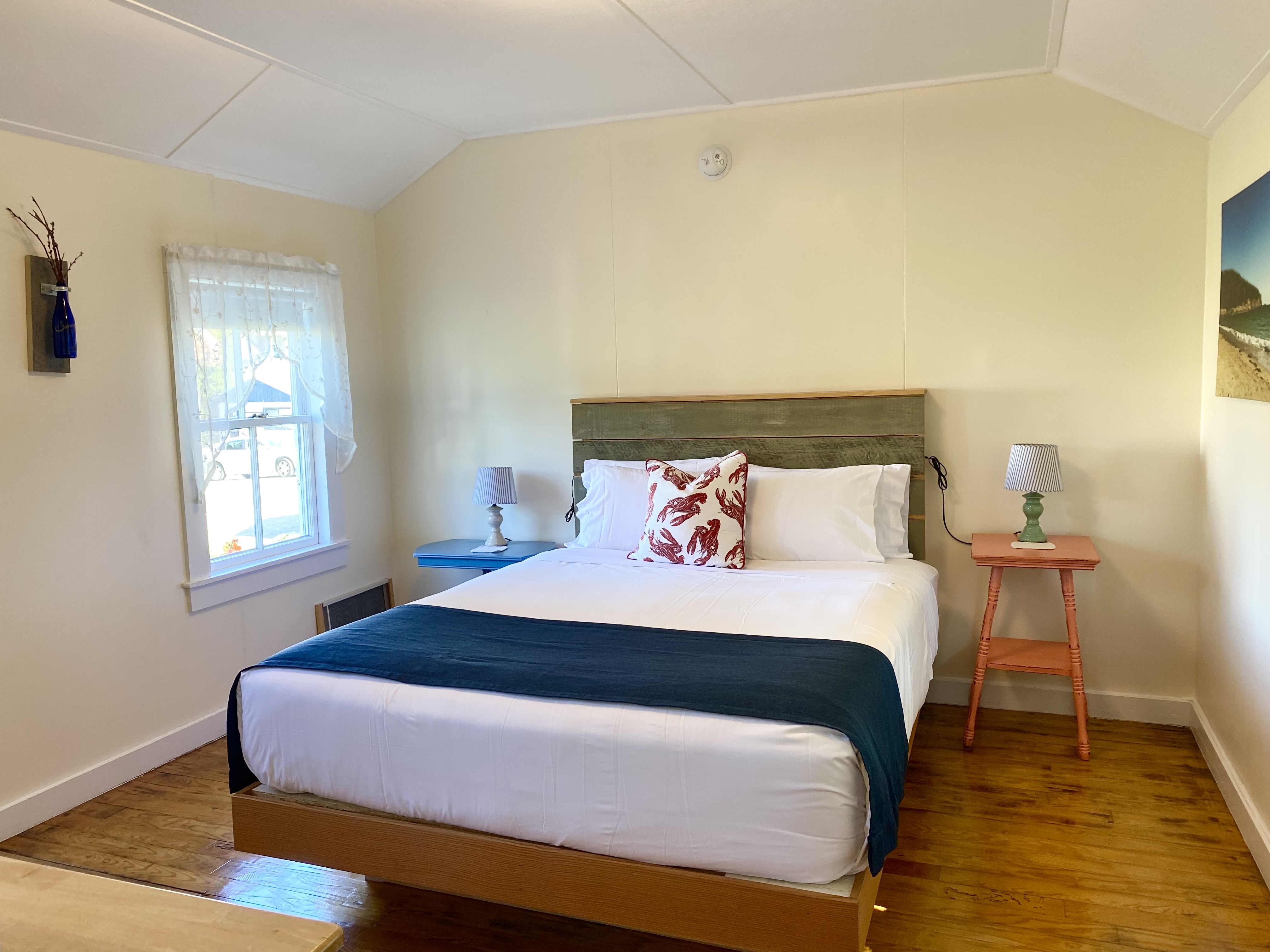 A neatly made bed with white sheets and a blue blanket in a room with cream-colored walls and wood floors.
