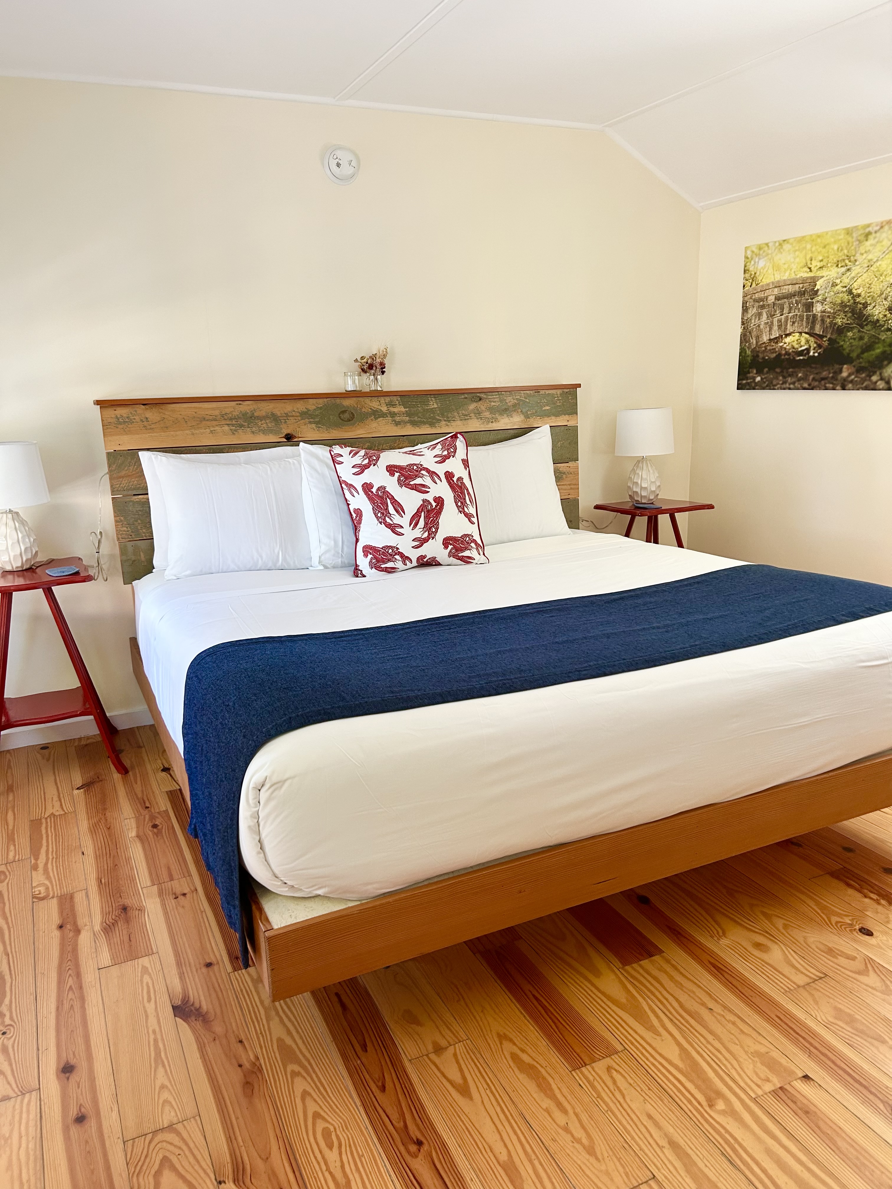 A neatly made bed with white bedding, a blue blanket, and red nightstands with lamps in a room with a wooden floor.