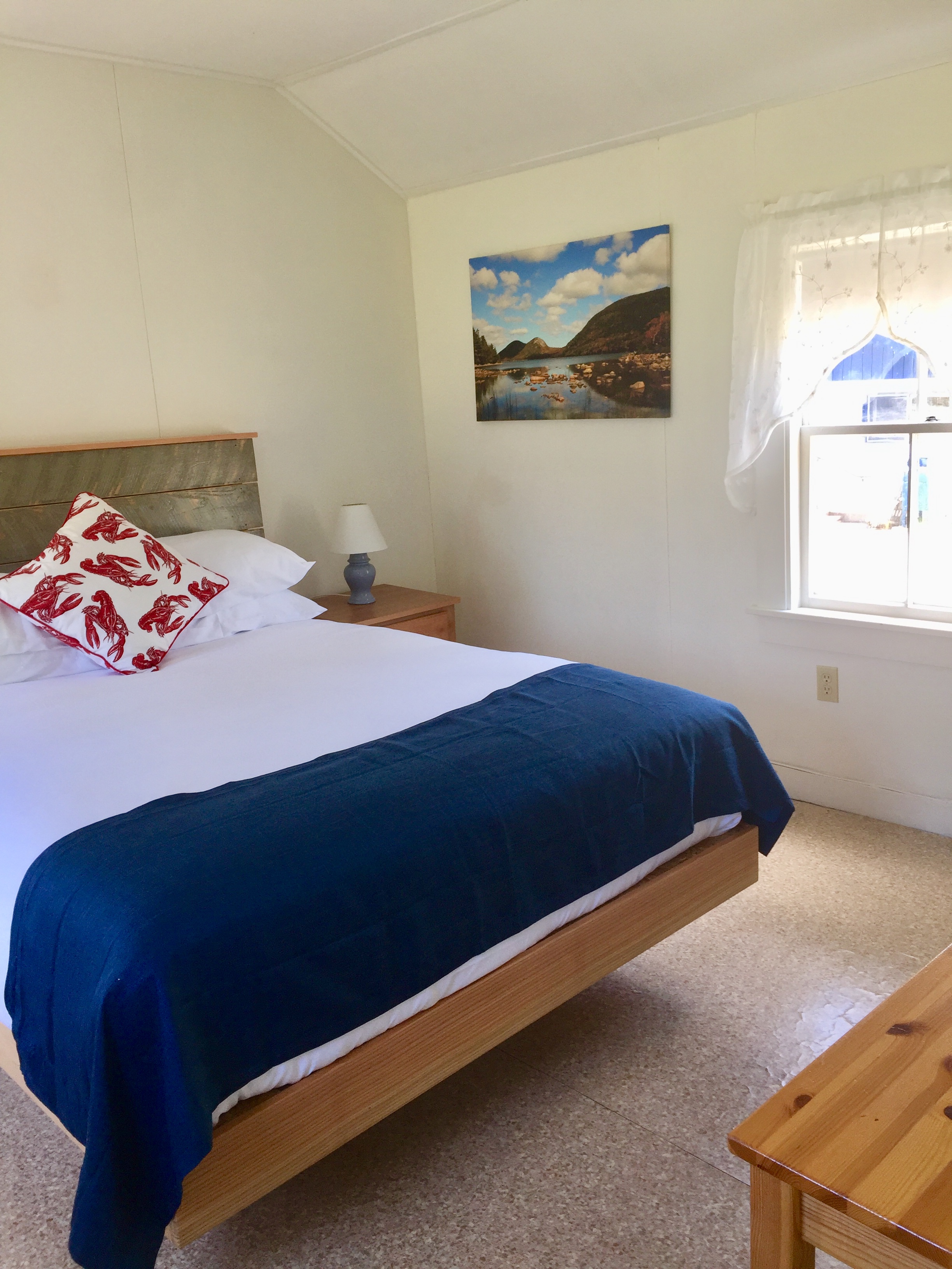 A neatly made bed with a blue blanket and white sheets, a nightstand with a lamp, and a window with a view in a cozy bedroom.