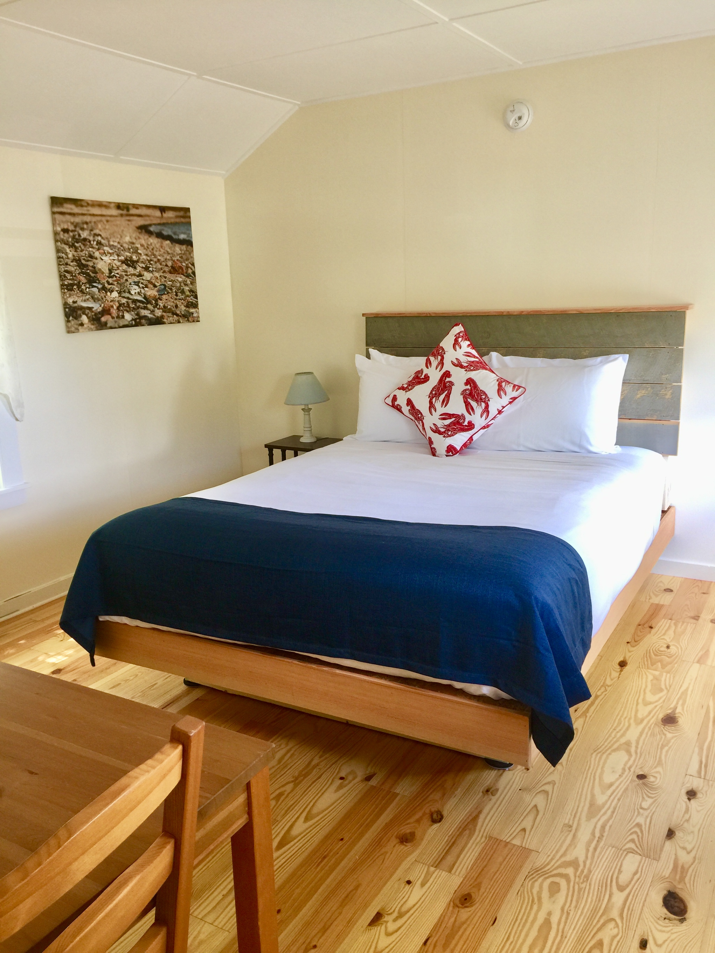 A neatly made bed with white sheets and a blue blanket, accompanied by a wooden table and chair in a cozy room with light wood flooring.