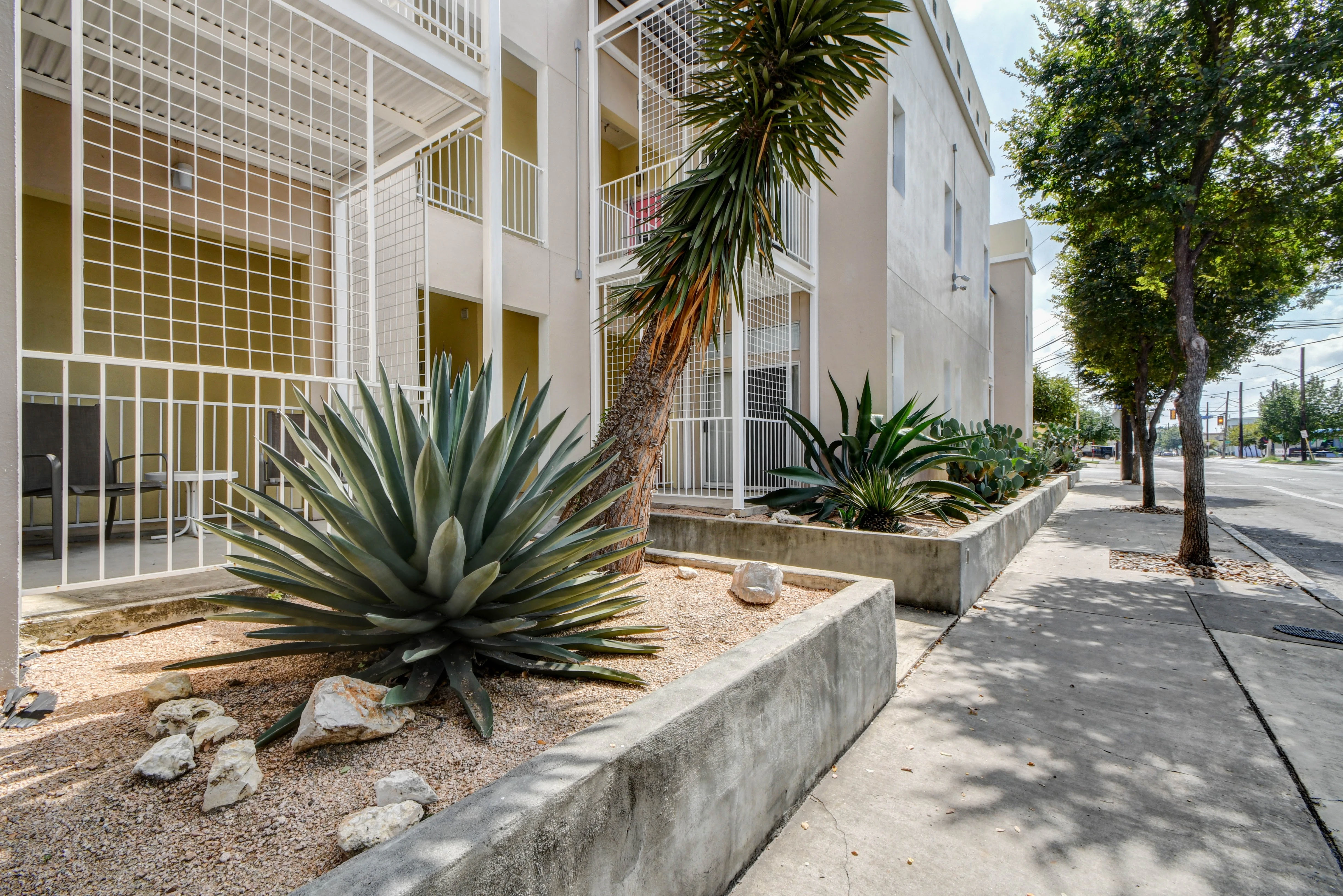 A beige building with white railings and balconies is seen from the street, with plants and trees in front of it.