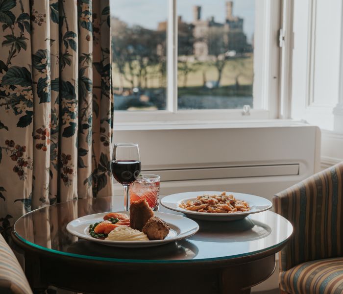 A table set with two plates of food and glasses of wine, placed in front of a window with views over the marina to castle.
