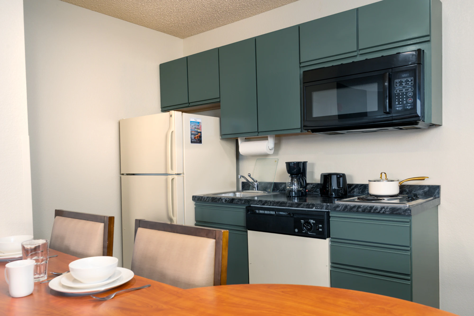 A kitchenette with green cabinets, black marble countertop, white refrigerator, and a dining table set for breakfast.