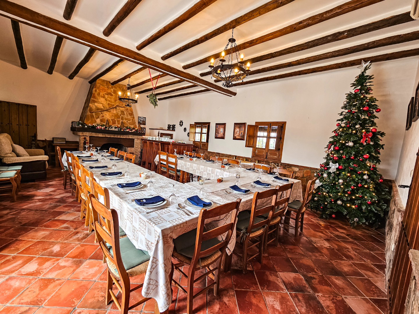 A rustic dining room with long tables set for a meal, featuring white tablecloths, neatly arranged plates, glasses,