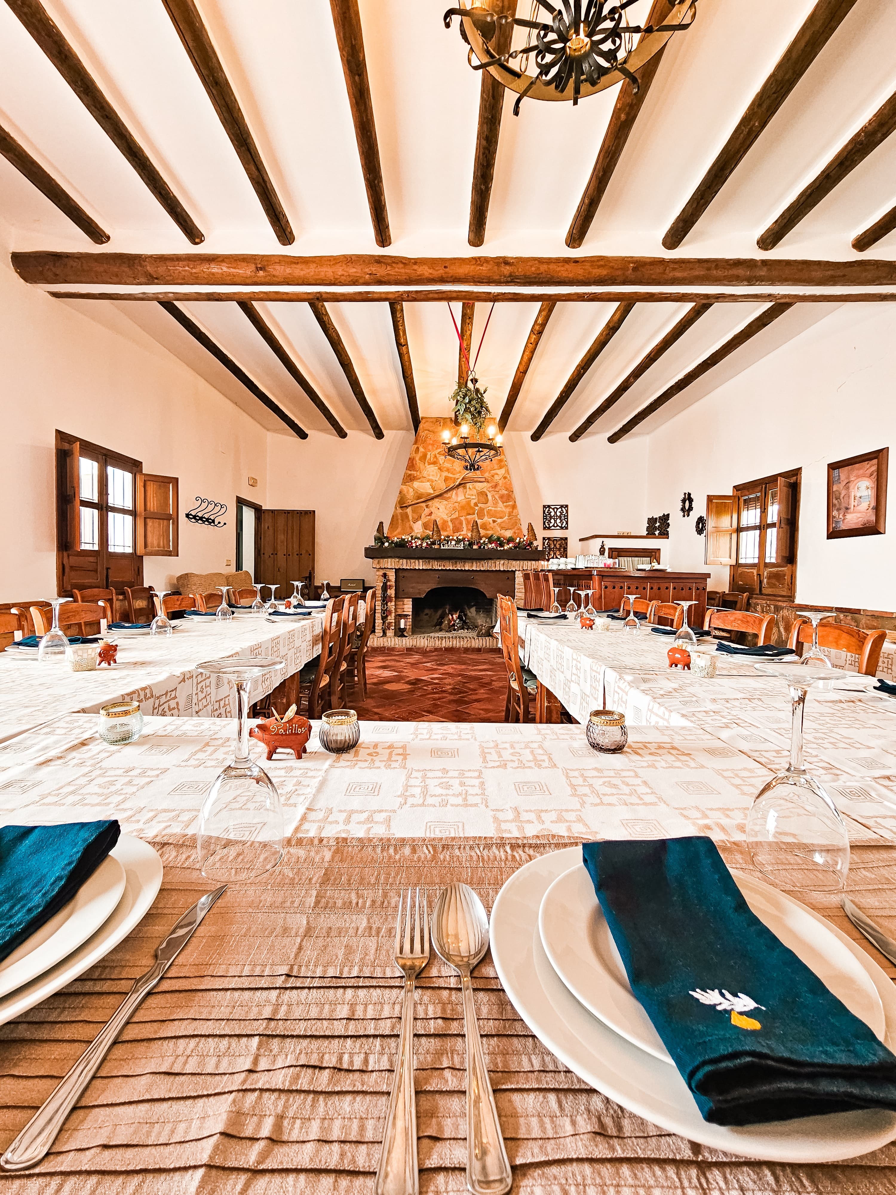 A rustic dining room with long tables set for a meal, featuring white tablecloths, neatly arranged plates, glasses,