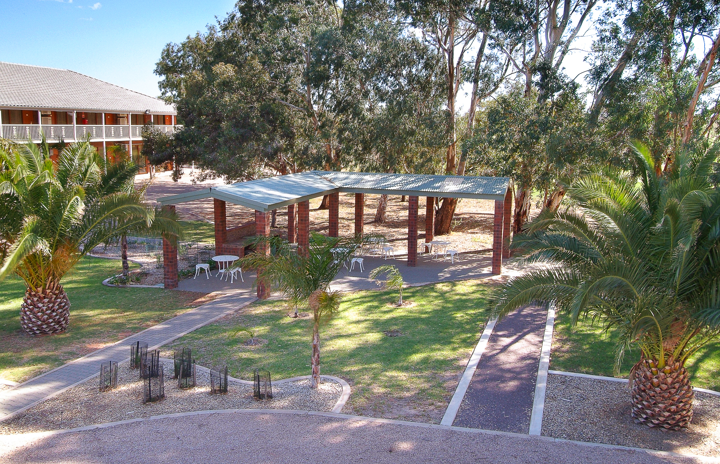 Lush green space with a covered picnic area, palm trees, and a multi-story building on the left.