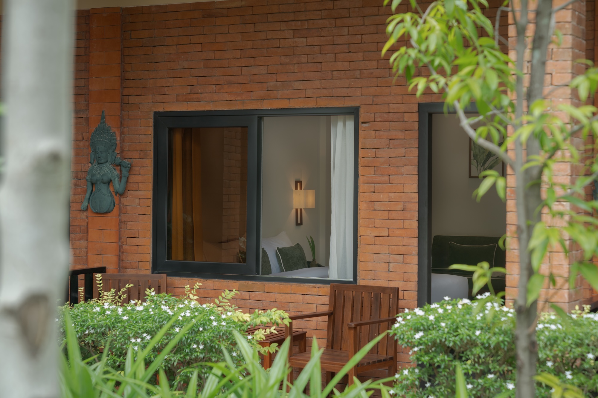 Exterior of hotel room with brick walls, open wooden shutters, bed, and lamp visible amid greenery