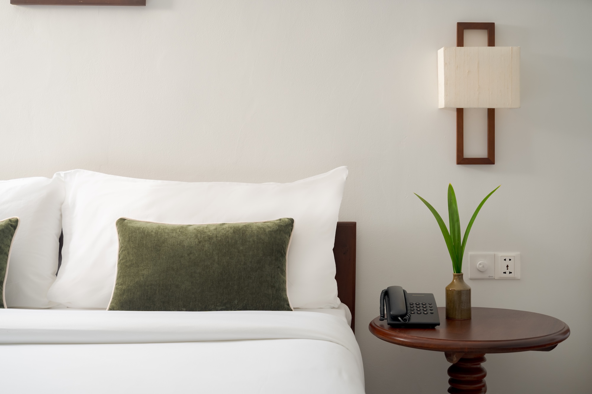 Neatly made bed with white linens, green pillow, wooden nightstand, lamp, and plant