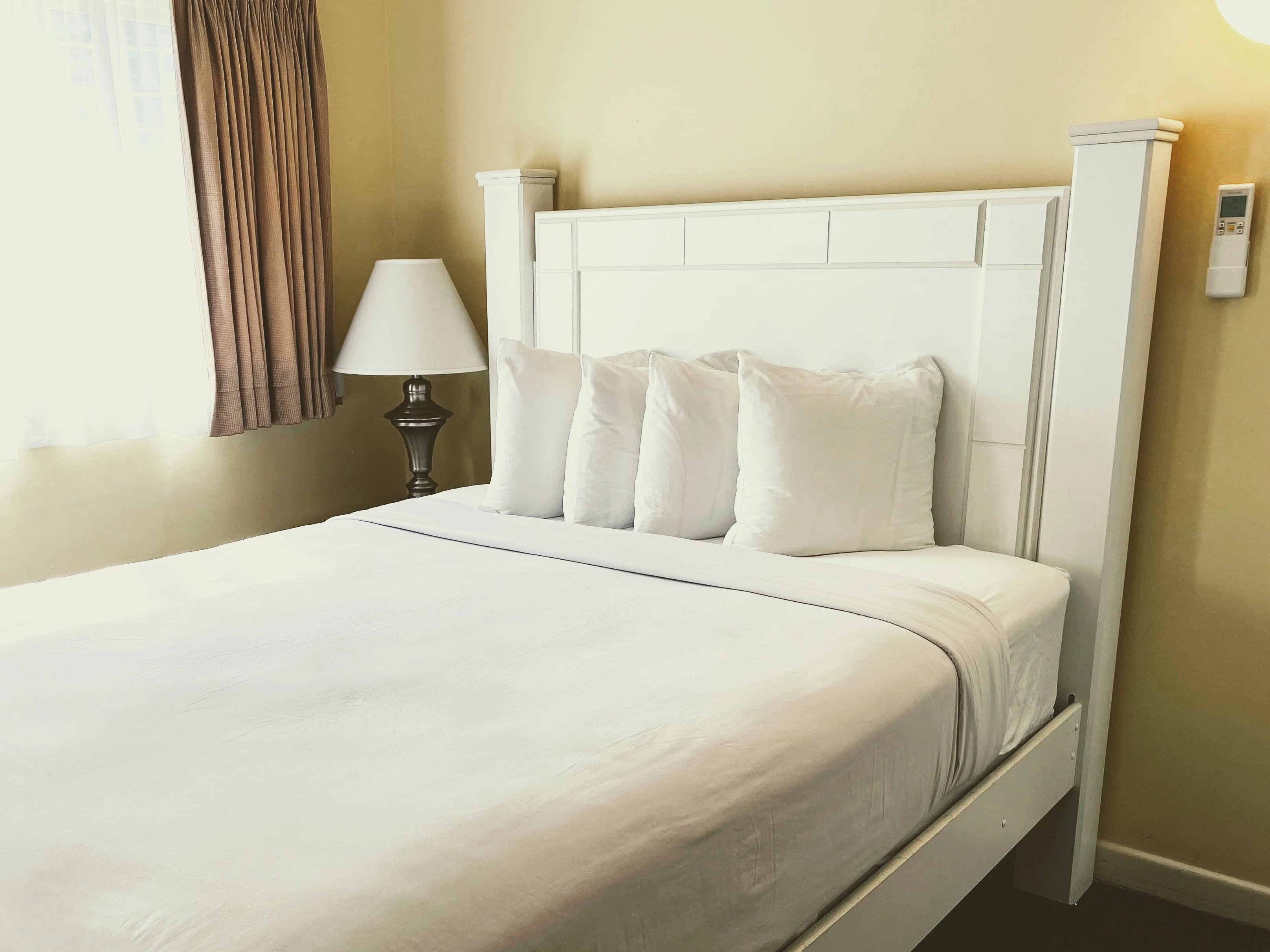 A neatly made bed with white linens and pillows, a bedside lamp, and a window with curtains.