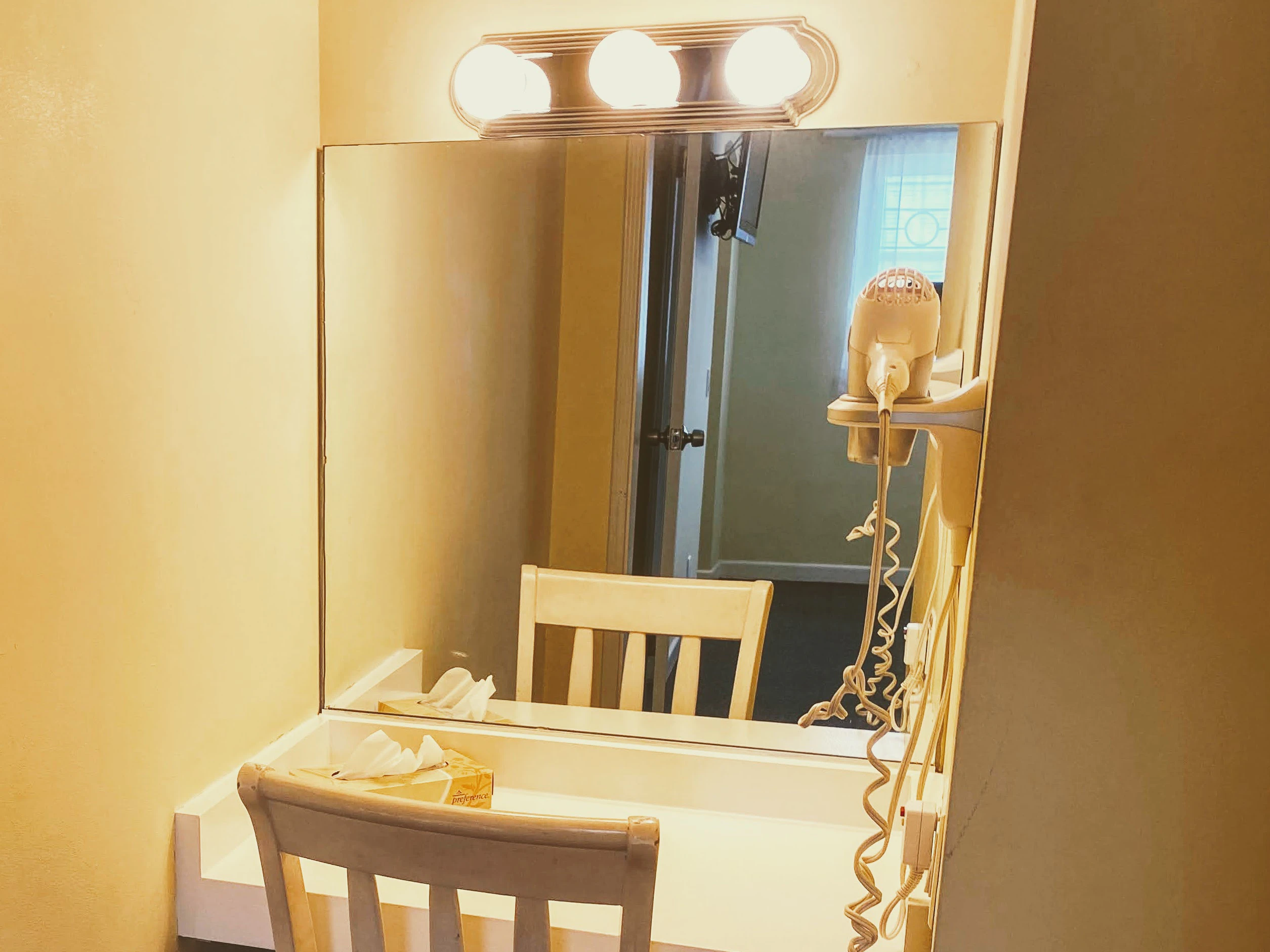 A bathroom vanity with a large mirror, lighting, a chair, and tissues.