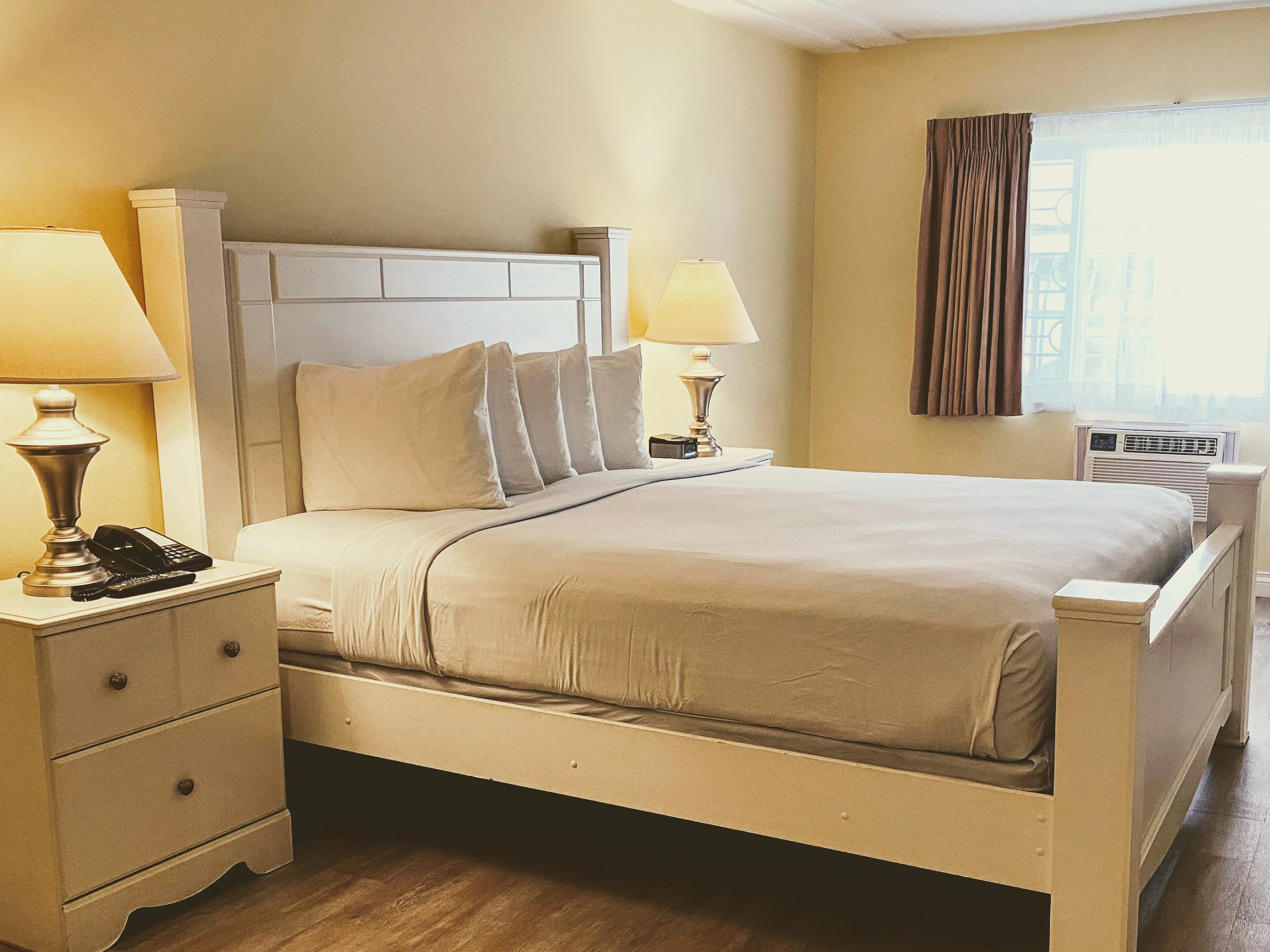 A large bed with white linens and pillows, two nightstands with lamps, and a window with curtains in a room with yellow walls and wood floors.